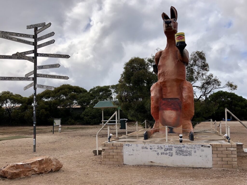 A kangaroo holding a Vegemite jar and distance signs to locations around the world. Border Village, SA/WA border.